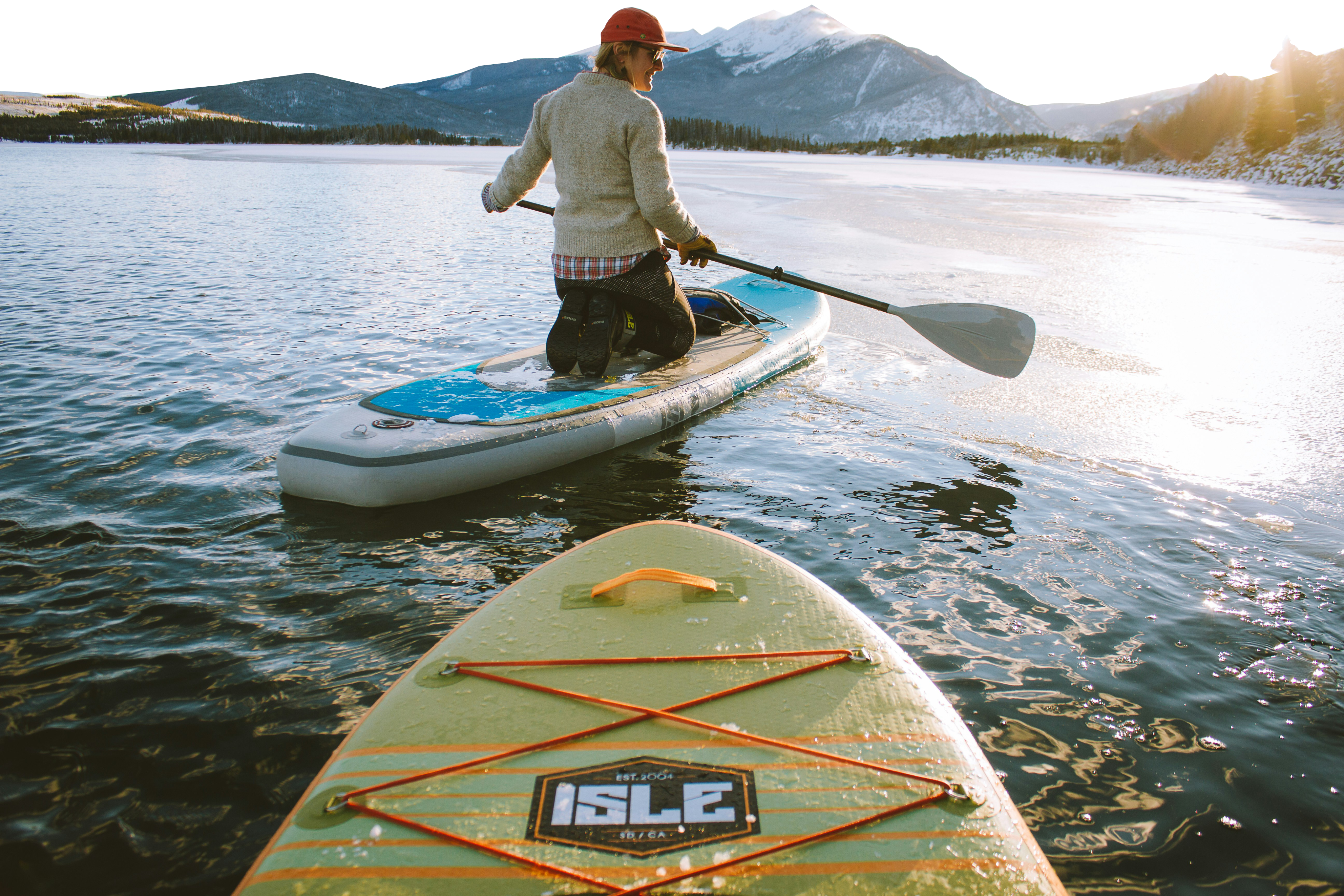 A person in a red hat and sweater kneels on a paddleboard, paddling across a calm lake surrounded by snowy mountains, with another paddleboard visible in the foreground.