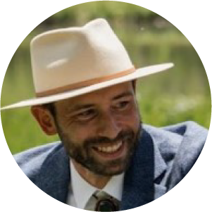 A man with a beard, wearing a light-colored fedora and blue blazer, smiles while looking to the side. The background is green and slightly blurred, suggesting an outdoor setting.
