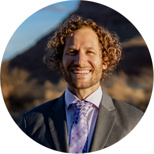 A man with curly hair wearing a gray suit, light pink shirt, and patterned tie smiles outdoors with blurred brown hills and blue sky in the background.