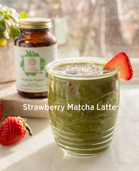 A glass jar filled with green strawberry matcha latte, topped with a strawberry slice. A jar of matcha powder is in the background, along with fresh strawberries. Sunlight creates a warm, inviting atmosphere.