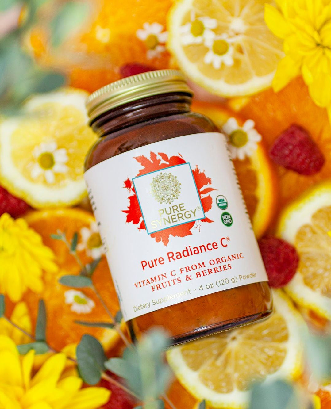 A bottle of Pure Synergy Pure Radiance C powder rests among vibrant sliced citrus fruits, raspberries, and yellow flowers. The bottle label highlights organic vitamin C from fruits and berries.