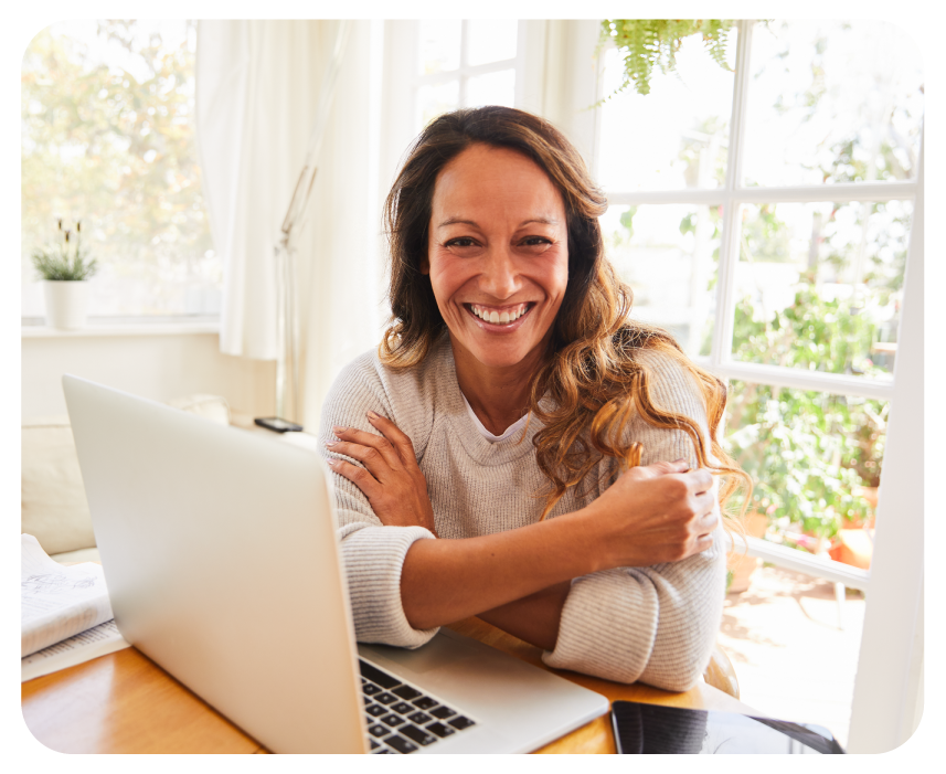 A woman with long brown hair smiles warmly while sitting at a table with a laptop in a bright, sunlit room filled with plants.