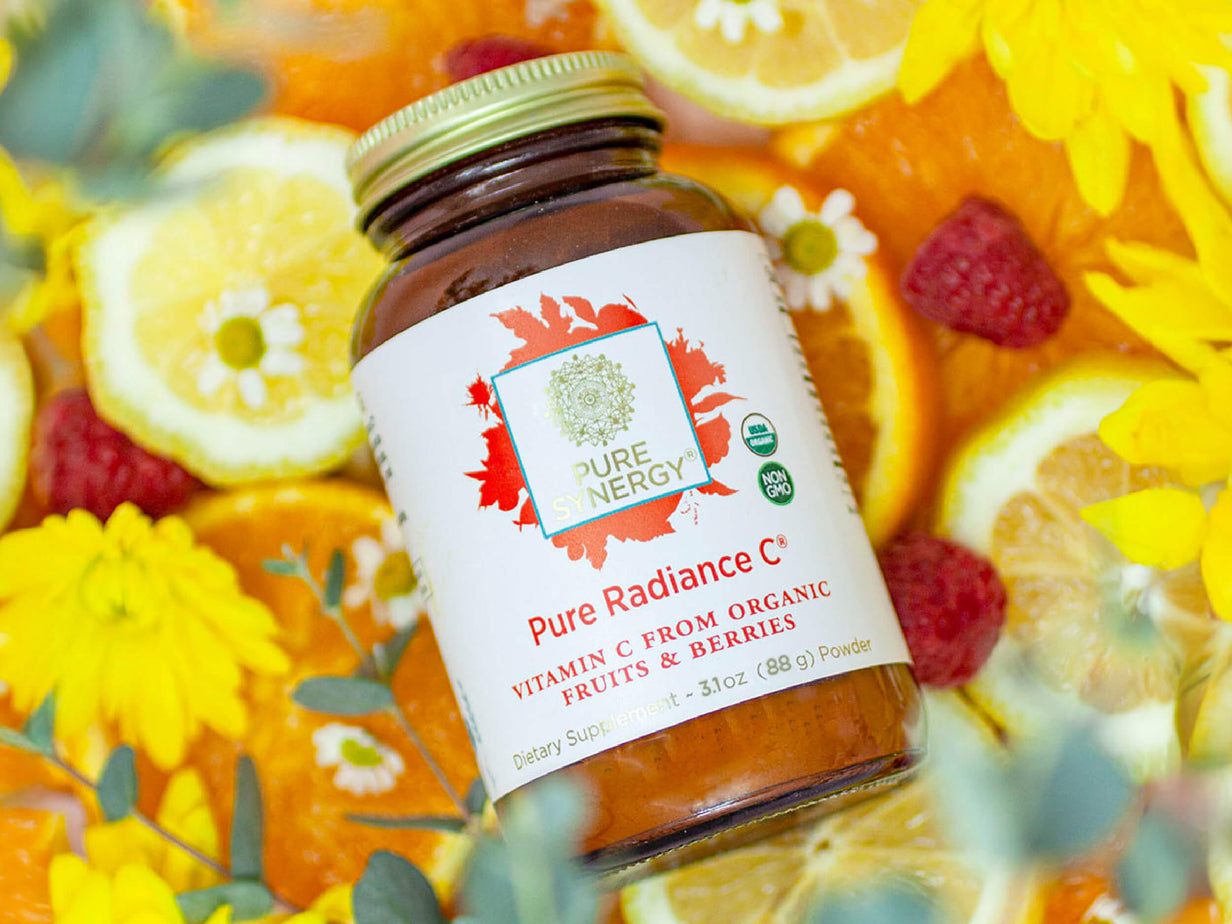 A bottle of Pure Synergy Pure Radiance C powder sits surrounded by sliced citrus fruits, raspberries, and yellow flowers.
