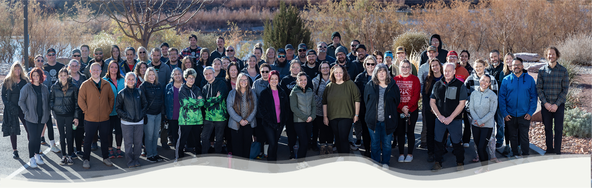 A group photo of about 80 people standing outdoors, posing together on a pathway. They are surrounded by trees with autumn foliage. The sky is clear, and everyone is dressed in casual, warm clothing.