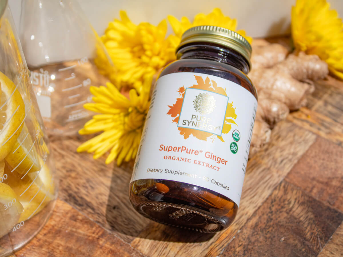 A bottle of Pure Synergy SuperPure Ginger organic extract capsules on a wooden surface, surrounded by bright yellow flowers, ginger root, a glass container, and lemon slices.