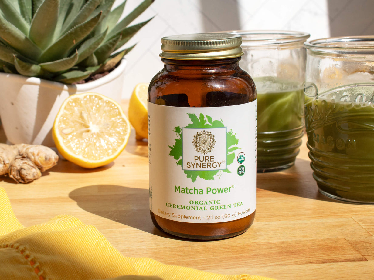 A jar of Pure Synergy Matcha Power organic green tea sits on a wooden surface. Next to it are a sliced lemon, ginger, an aloe plant, and two jars of green liquid. Sunlight casts shadows across the scene.