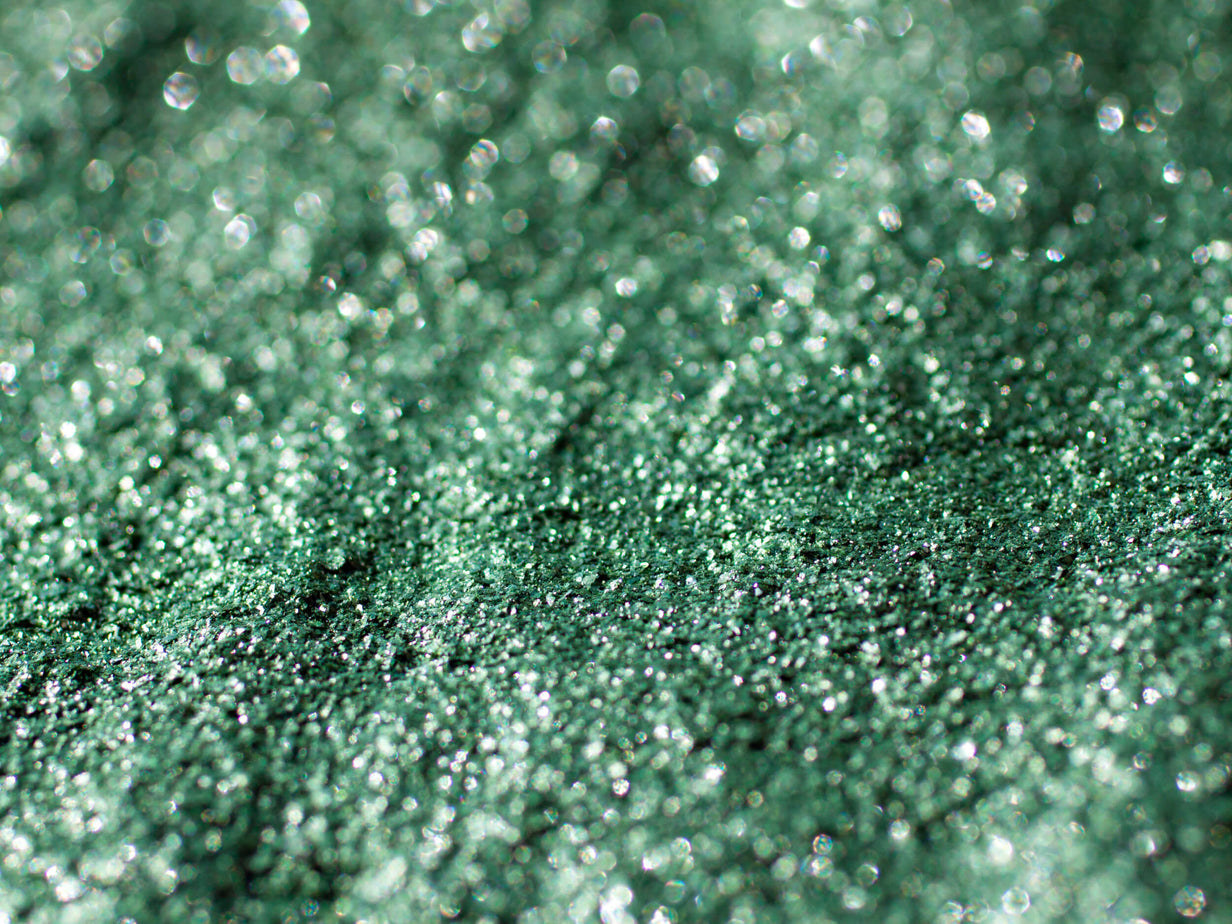 Close-up of a sparkling green textured surface, with a bokeh effect creating a shimmering appearance. The image focuses on the small, glittering particles reflecting light, giving an impression of a glistening, abstract landscape.