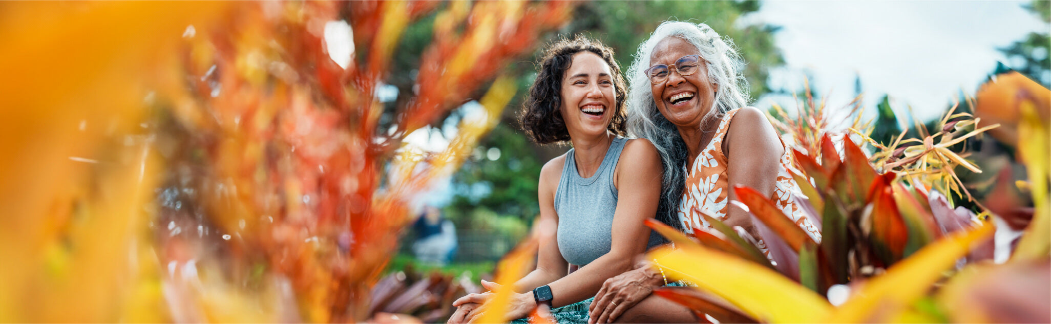 Two women, one younger and one older, sit laughing among vibrant tropical plants. The woman on the left has curly hair and wears a sleeveless top, while the woman on the right has gray hair and glasses, wearing a floral dress. Bright foliage surrounds them.