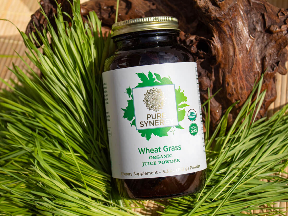 A jar of Pure Synergy Wheat Grass Organic Juice Powder is displayed on green grass, with a backdrop of a wooden texture. The label features green leaves and organic certification badges.