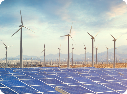 Rows of solar panels in the foreground with multiple wind turbines in the background, set in a sunny, open landscape with mountains in the distance.